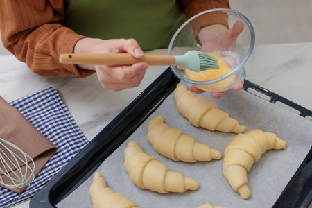 Woman brushing egg wash onto raw croissants at white marble table, closeup