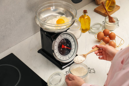 Woman holding spoon with flour near mechanical kitchen scale at white marble countertop indoors,...