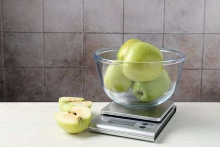 Electronic kitchen scale with bowl of green apples on white wooden table against tiled wallの写真素材