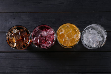 Refreshing soda water of different flavors with ice cubes in glasses on black wooden table, flat layの写真素材