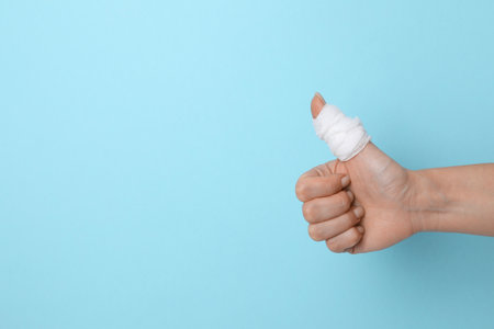 Woman with medical bandage on finger showing thumbs up against light blue background, closeup. Space for textの写真素材