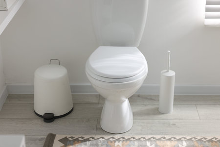 Toilet, brush and trash bin near white wall in bathroom