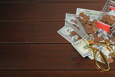 Dollar banknotes, scissors, coins and small shopping cart on wooden table, space for text