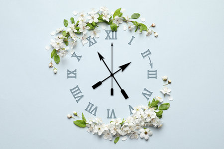 Clock and white flowers on light blue background, top view. Arrow showing time change one hour ahead in spring. Daylight saving timeの写真素材