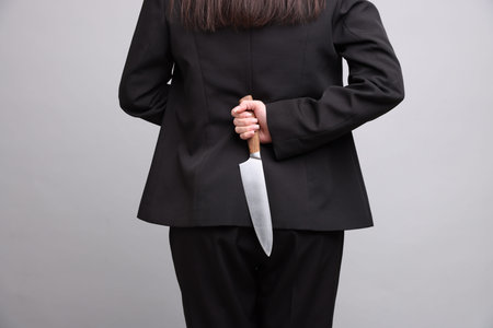 Businesswoman with knife behind her back on light gray background, closeup