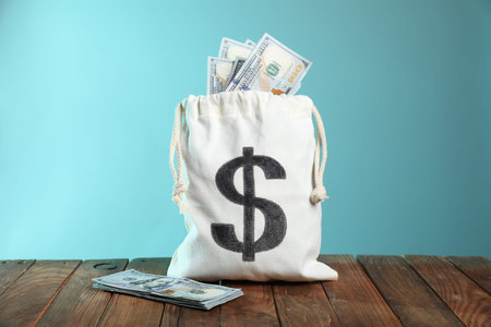 Burlap sack with dollar banknotes on wooden table against light blue background