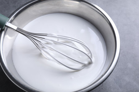 Whisk and whipped cream in bowl on gray table, closeup