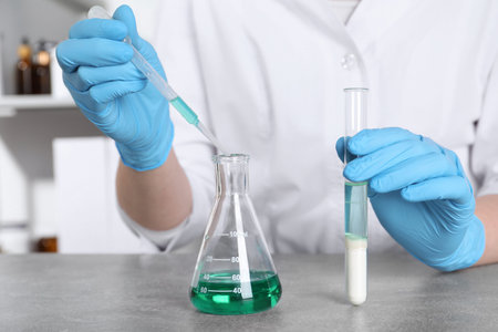 Laboratory testing. Scientist with test tube taking sample from flask at grey table indoors, closeup