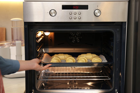 Woman putting baking sheet with raw croissants into oven indoors, closeup