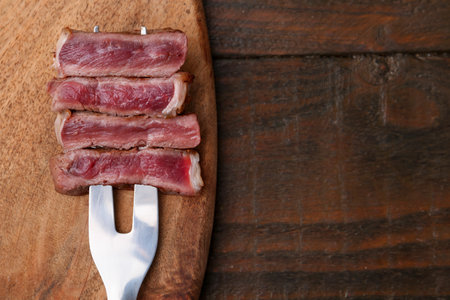 Carving fork with pieces of delicious beef on wooden table, top view. Space for textの写真素材
