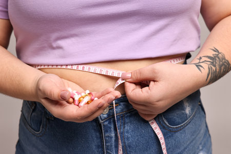 Plus size woman measuring waist with tape and holding pile of weight loss supplements on grey background, closeupの写真素材
