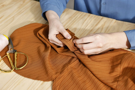 Woman sewing brown shirt with needle and thread at wooden table, closeupの写真素材