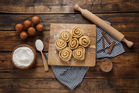 Raw cinnamon rolls and different ingredients on wooden table, flat lay