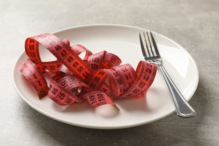 Bulimia. Fork, plate and measuring tape on grey table, closeup