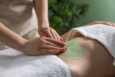 Esthetician applying cosmetic product for body wraps treatment onto woman's belly in spa salon, closeupの写真素材