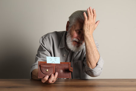 Upset senior man holding wallet with drawn sad face at wooden table. Financial problemsの写真素材