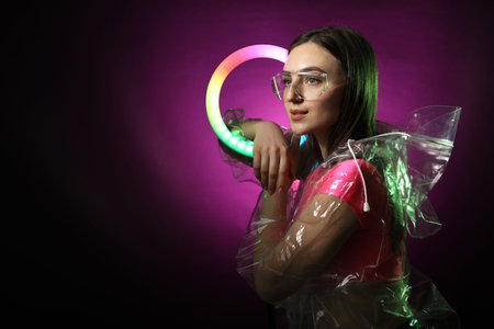 Stylish woman with ring lamp wearing transparent coat and glasses on dark purple background, space...