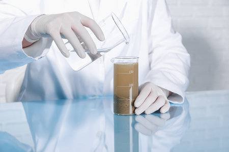 Laboratory testing. Scientist pouring liquid into beaker with soil sample at table indoors, closeup