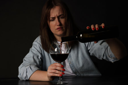 Alcohol addiction. Miserable woman pouring wine into glass at table in darkの写真素材
