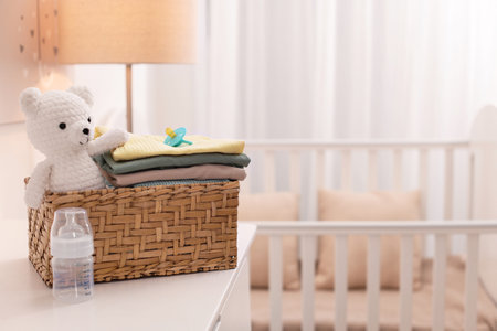 Wicker basket with child's clothes, toy bear and feeding bottle on chest of drawers indoors. Space...