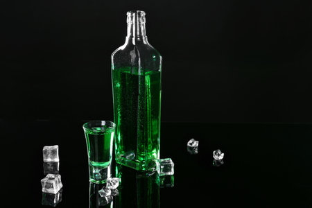 Absinthe in shot glass, bottle and ice cubes on black mirror surface, space for textの写真素材