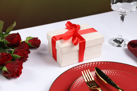 Gift box, plates, cutlery and roses on white table against black background, closeup. Table setting for romantic dinnerの写真素材