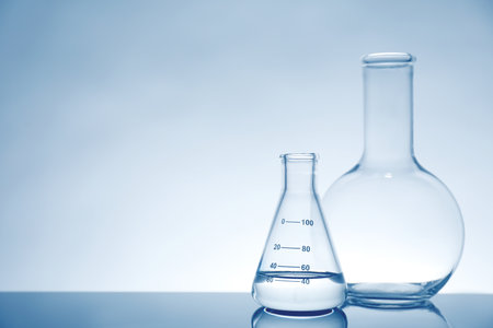 Laboratory glassware with liquid on mirror surface against light background, space for textの写真素材