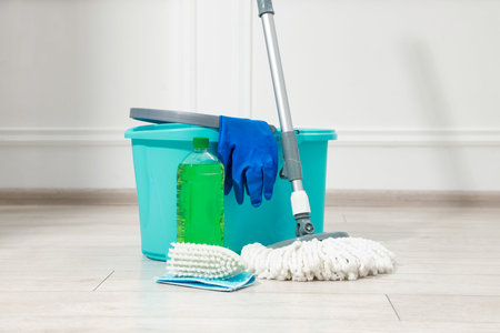 String mop, brush, detergent, bucket and gloves on wooden floor near white wall