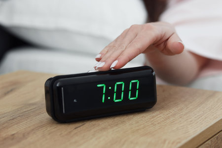 Woman turning off digital alarm clock on bedside table indoors, closeup