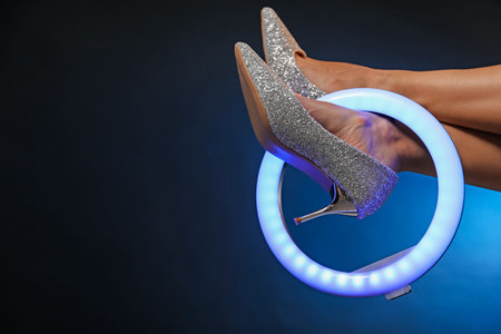 Woman with ring lamp wearing high heeled shoes on dark blue background, closeup. Space for textの写真素材