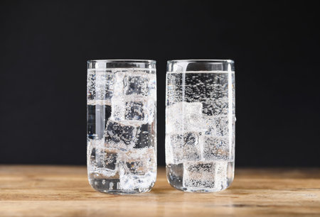 Refreshing soda water with ice cubes in glasses on wooden table against black background, closeupの写真素材