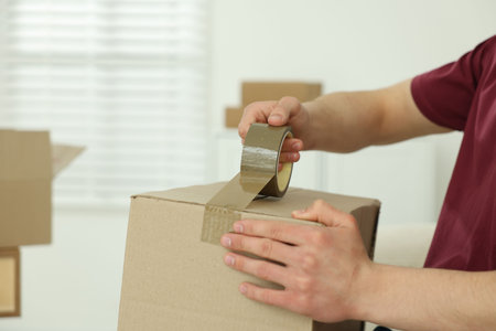 Man sealing cardboard box with adhesive tape indoors, closeupの写真素材