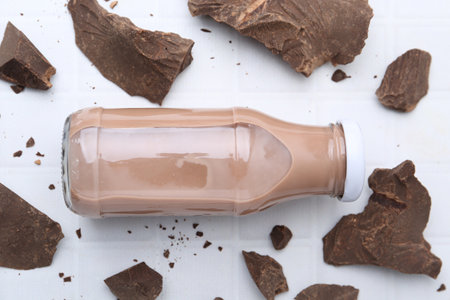 Tasty chocolate milk in glass bottle and pieces of chocolate on white table, flat layの写真素材