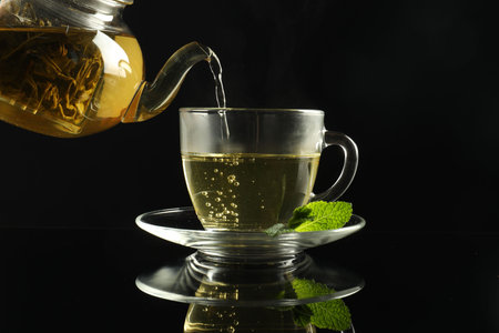 Pouring freshly brewed tea from teapot into cup at dark mirror table, closeupの写真素材
