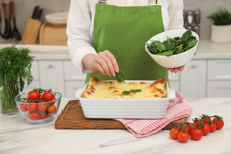 Woman putting spinach onto lasagna at white marble table in kitchen, closeupの写真素材