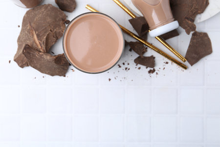Tasty chocolate milk in glass, bottle and pieces of chocolate on white tiled table, flat lay. Space for textの写真素材