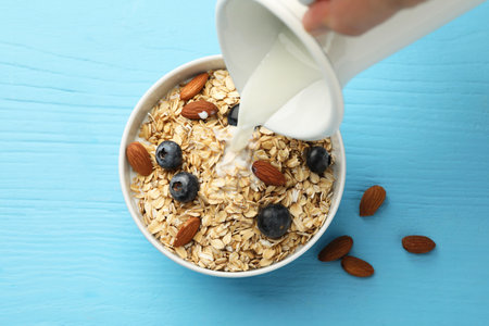 Woman pouring milk into bowl with oatmeal, blueberries and almonds at light blue table, topの写真素材