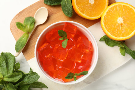 Delicious jelly cubes in bowl, mint, spoon and orange on white marble table, flat layの写真素材
