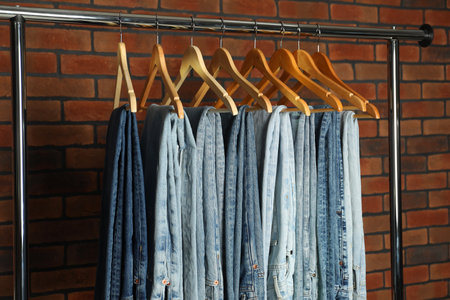 Many stylish jeans hanging from rack near brick wallの写真素材