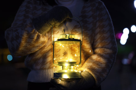 Woman holding Christmas lantern with burning candle and fairy lights in darkness, closeupの写真素材