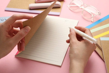 Woman writing in copybook on pink background, closeup