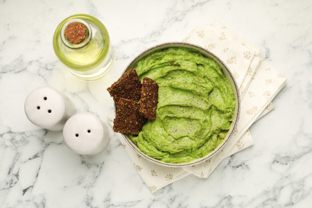 Tasty avocado dip in bowl, crispbreads and oil on white marble table, flat lay