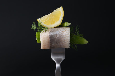 Fork with marinated herring fillet, cucumber, dill and lemon on black background, closeupの写真素材