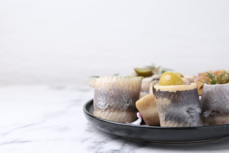 Delicious herring rolls with olives, pickles and dill on light marble table, closeup. Space for text