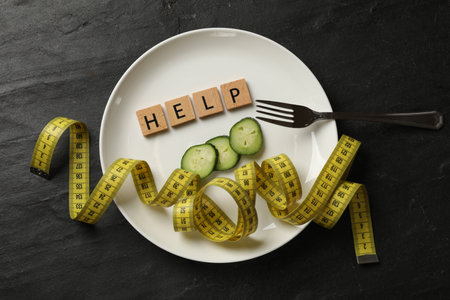Plate with word Help, pieces of cucumber, measuring tape and fork on dark textured background, top viewの写真素材
