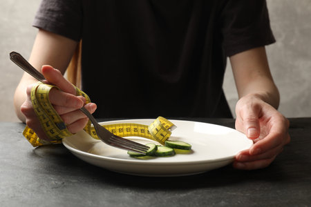 Woman with measuring tape eating pieces of cucumber at dark textured table, closeupの写真素材