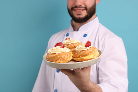 Happy confectioner in uniform holding delicious profiteroles with strawberries on light blue...