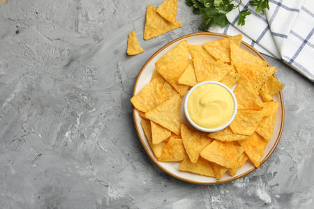 Tasty cheese dipping sauce in bowl, nacho chips and parsley on grey table, flat lay. Space for textの写真素材