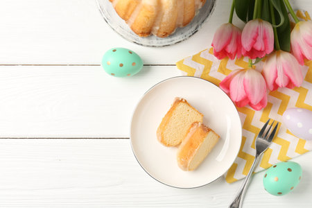 Pieces of delicious bundt cake, Easter eggs and tulips on white wooden table, flat lay. Space for...