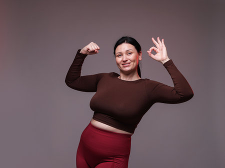 Plus size woman in gym clothes showing ok gesture on grey backgroundの写真素材